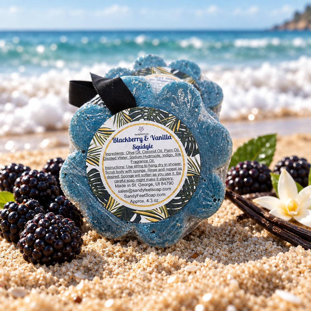 A packaged blue flower-shaped soap in a sponge with a black ribbon and a round label reading 'Blackberry & Vanilla Squdgie' by Sweetful Soap, photographed on a plain light background.